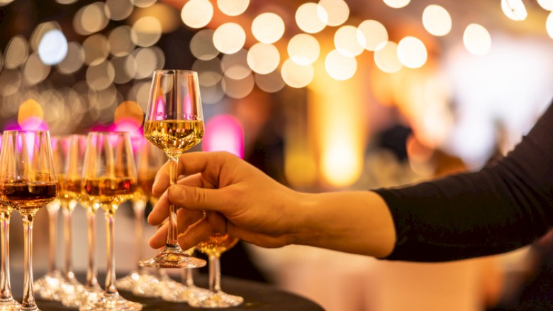 A person is holding a glass of white wine at an event or celebration with multiple glasses and warm, bokeh lights in the background.