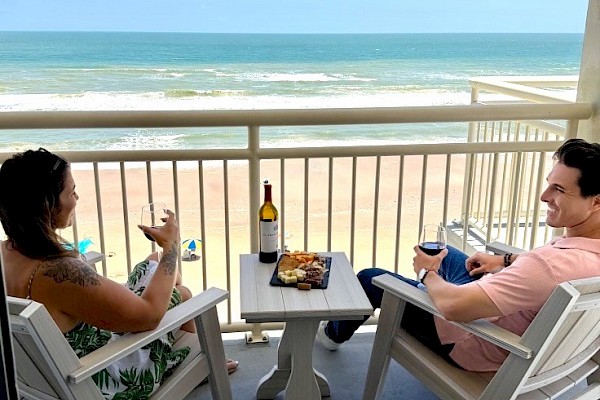 A couple enjoys wine and snacks on a balcony overlooking a sunny beach and the ocean in this picturesque seaside setting.