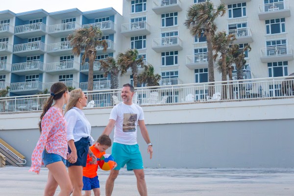 A group of people is walking on a sandy beach, with a modern multi-story building and palm trees in the background.