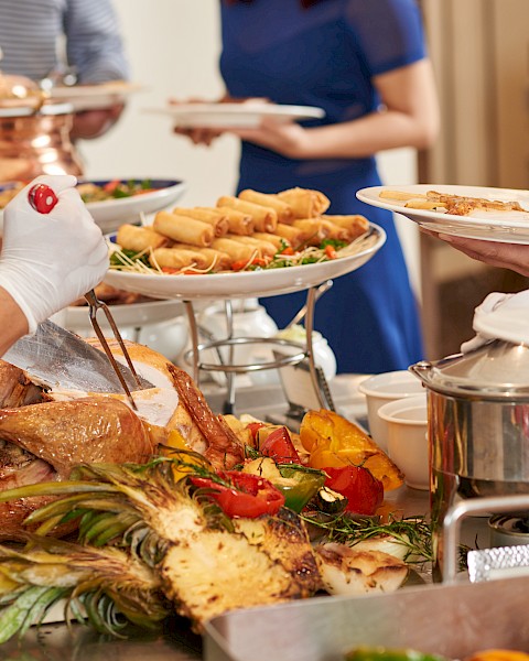 A buffet with carved meat, grilled vegetables, spring rolls, and people serving themselves from a selection of dishes.