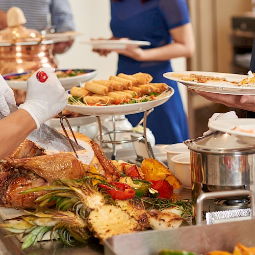 A buffet with carved meat, grilled vegetables, spring rolls, and people serving themselves from a selection of dishes.