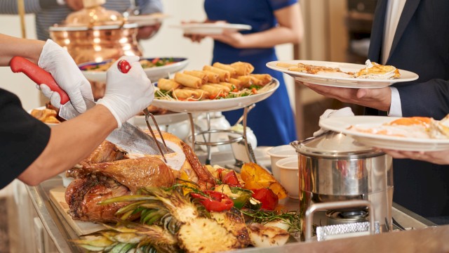 A buffet with carved meat, grilled vegetables, spring rolls, and people serving themselves from a selection of dishes.