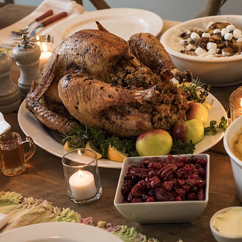 A festive dinner spread with a roasted turkey centerpiece, sides like stuffing, cranberry sauce, mashed dishes, candles, and desserts on a wooden table.