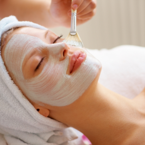 A person is relaxing with a facial mask being applied by a brush, wearing a towel wrapped around their head.