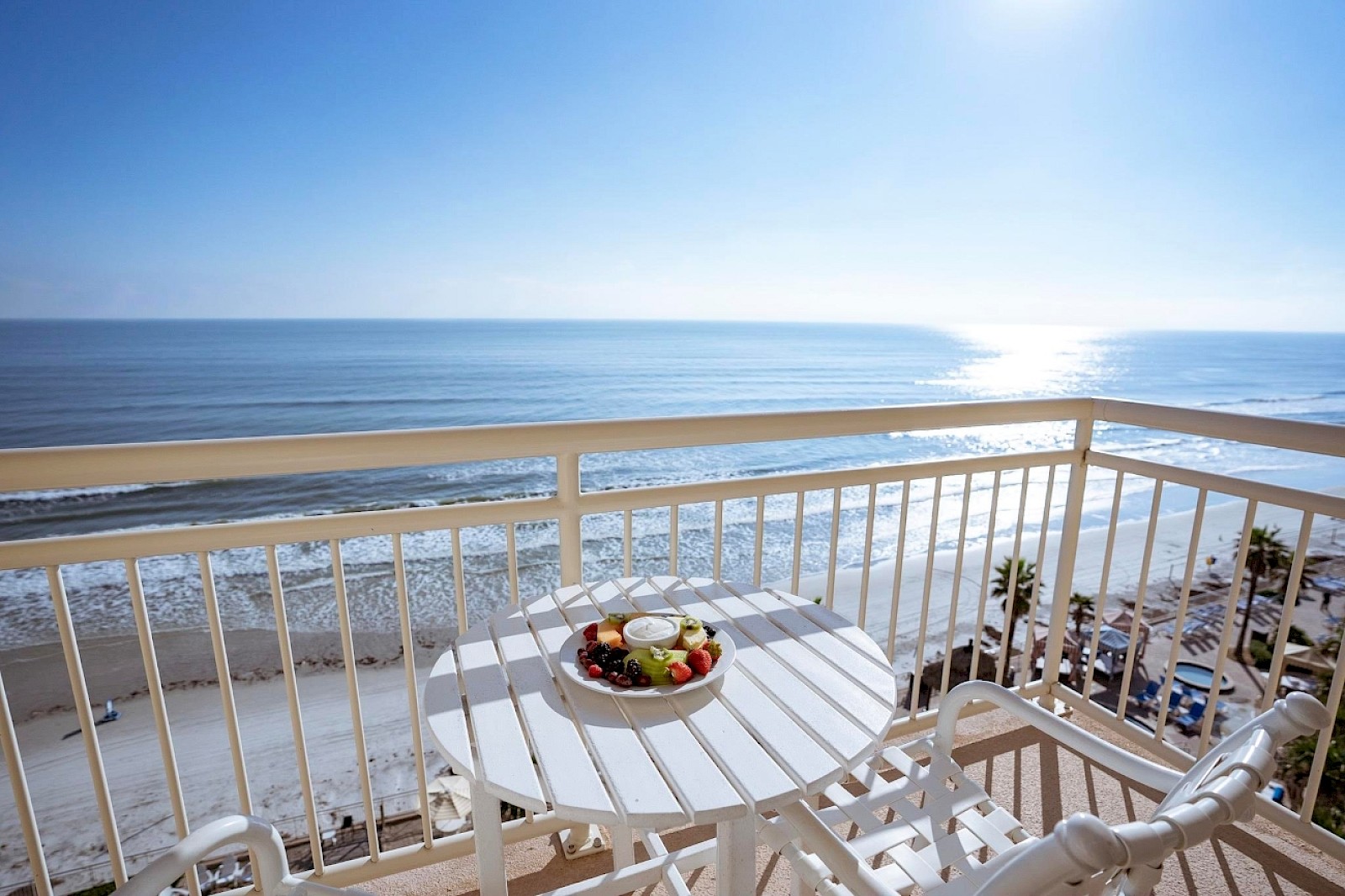 A white patio table with fruit and dip overlooks a sunny beach and ocean from a balcony.