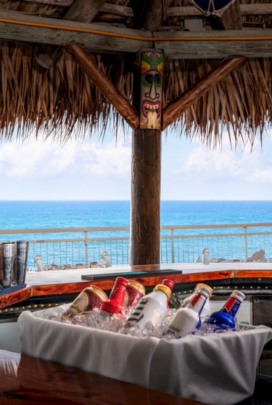 A tropical beach bar with a thatched roof, seaside view, and various bottles of alcohol on the counter.