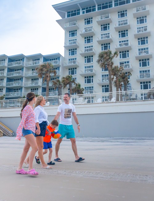 A family of four walks along a beachside path near a modern, multi-story hotel with palm trees and balconies.
