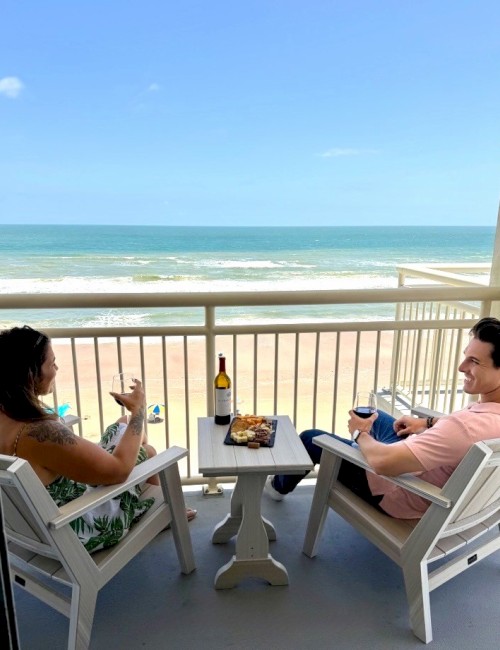 A couple enjoys wine and snacks on a balcony overlooking a beach and ocean under a clear blue sky.