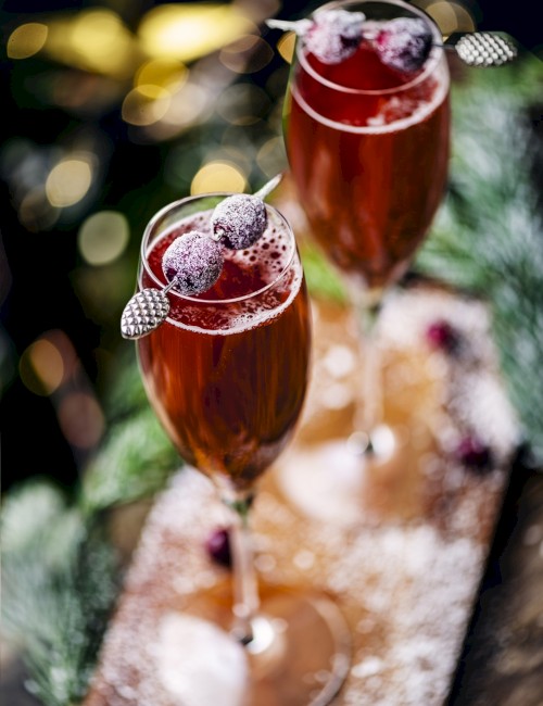 Two glasses of a red cocktail garnished with sugared cranberries on sticks, set on a table dusted with powdered sugar and festive decor.