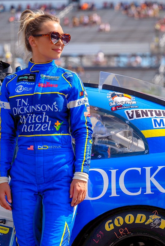 A woman in a blue racing suit stands beside a blue race car with various sponsor logos at a track, preparing for a racing event.