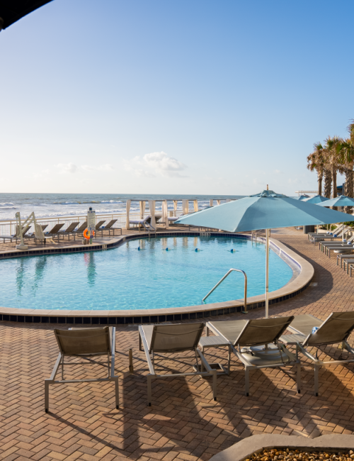 A serene beachside pool area with sun loungers, umbrellas, palm trees, and a view of the ocean and sky.