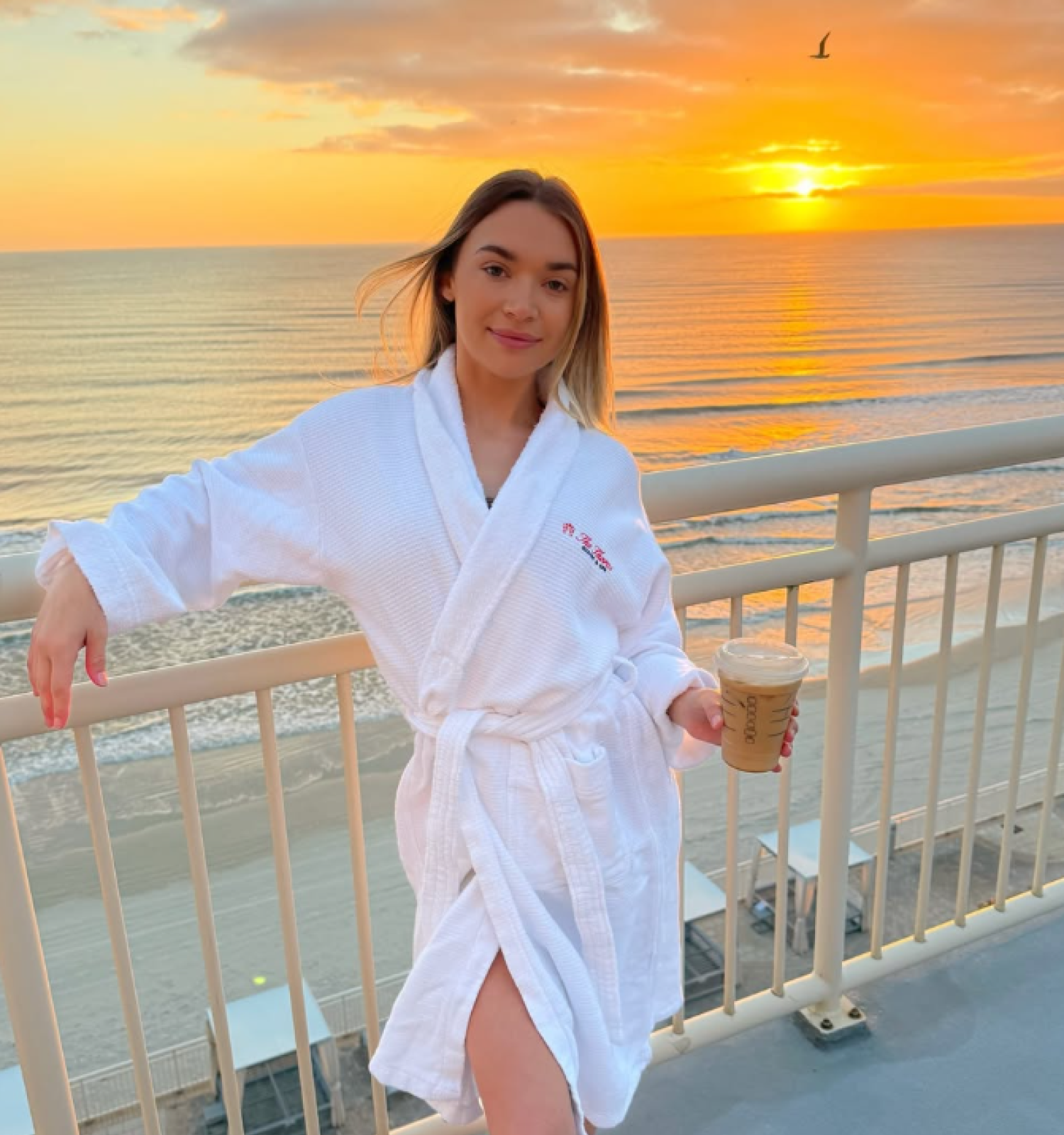 A woman in a white robe enjoying coffee on a balcony during a beautiful sunset over the ocean.