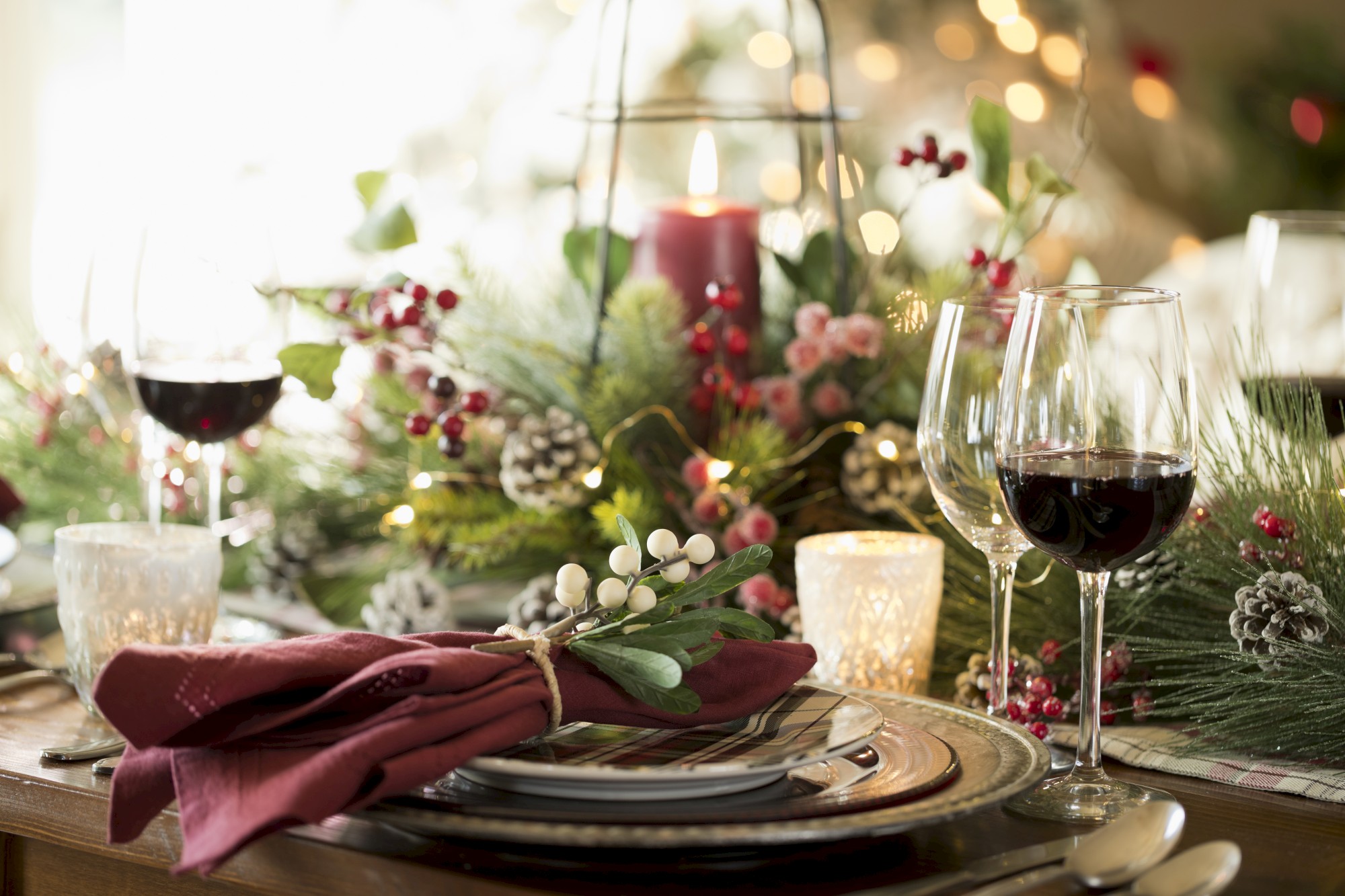 A festive table setting with wine glasses, candles, red napkins, and a centerpiece of greenery, berries, and pinecones.