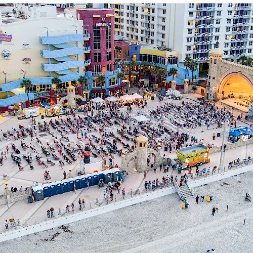 A crowded beach area near colorful buildings with many people sitting, walking, and enjoying time by the ocean.