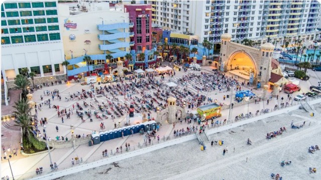 A crowded beach area near colorful buildings with many people sitting, walking, and enjoying time by the ocean.