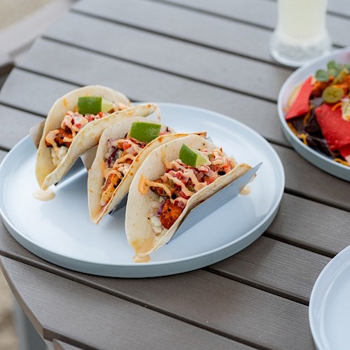 Three tacos filled with seasoned meat, cheese, and toppings, garnished with lime wedges on a white plate.