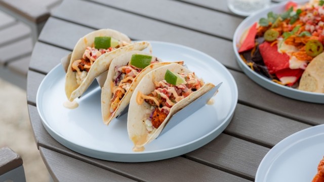 Three tacos filled with seasoned meat, cheese, and toppings, garnished with lime wedges on a white plate.