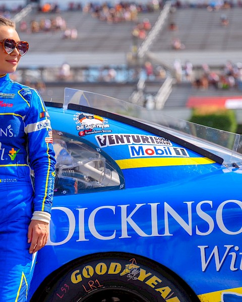 A female race car driver in a blue suit stands next to her blue racing car on the track, with stadium bleachers in the background.