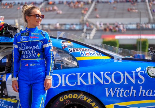 A female race car driver in a blue suit stands next to her blue racing car on the track, with stadium bleachers in the background.
