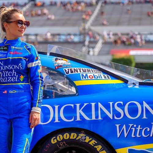 A female race car driver in a blue suit stands next to her blue racing car on the track, with stadium bleachers in the background.