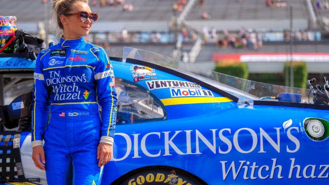 A female race car driver in a blue suit stands next to her blue racing car on the track, with stadium bleachers in the background.