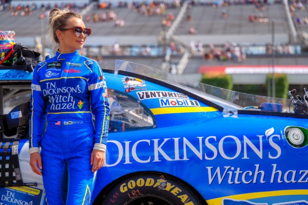 A female race car driver in blue racing suit standing next to a blue stock car on a racetrack with spectators in the background.