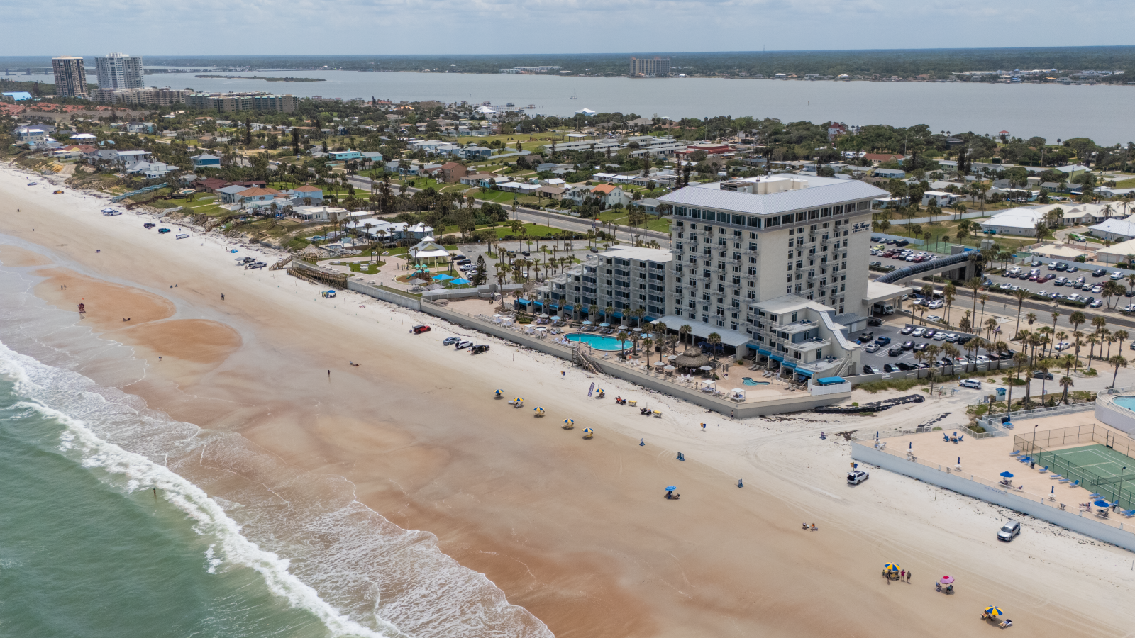 An aerial view of a beach resort with a large hotel, beach umbrellas, and a sandy shoreline along the ocean.
