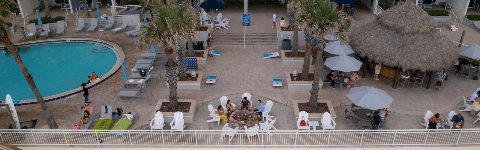 A beachfront hotel with a swimming pool, people relaxing on lounge chairs, a thatched umbrella area, and a staircase leading to the beach.