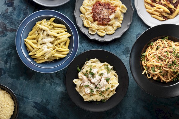 Assorted plates of pasta including penne, fettuccine, and rigatoni with various sauces and toppings on a dark teal surface.