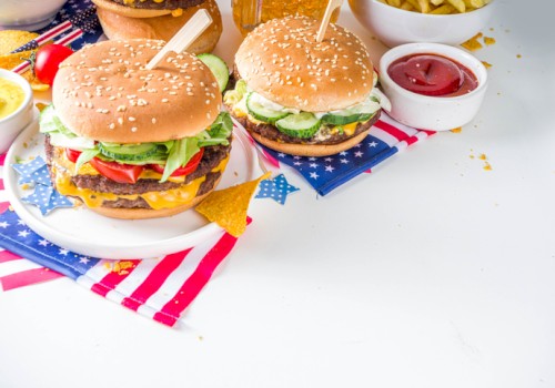 Dos hamburguesas con queso, lechuga, tomate y pepinillos en una mesa con motivos patrióticos, acompañadas de patatas fritas y kétchup.