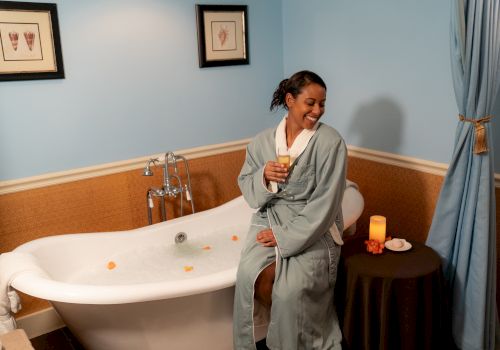 A person in a bathrobe sits on a tub edge with a drink, next to a table with a candle, in a cozy bathroom.