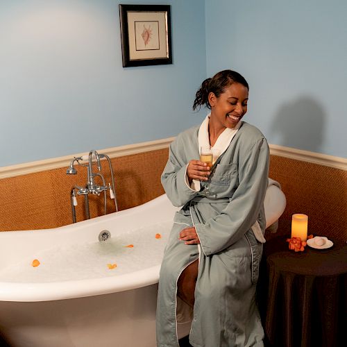 A person in a bathrobe sits on a tub edge with a drink, next to a table with a candle, in a cozy bathroom.