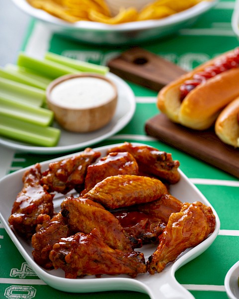 Buffalo chicken wings with celery sticks, ranch dipping sauce, potato chips, onion rings, hot dogs, and chocolate brownies on a football-themed table.