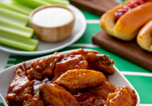 Buffalo chicken wings with celery sticks, ranch dipping sauce, potato chips, onion rings, hot dogs, and chocolate brownies on a football-themed table.