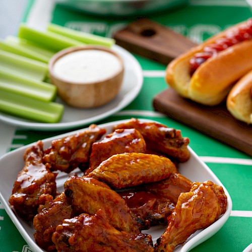 Buffalo chicken wings with celery sticks, ranch dipping sauce, potato chips, onion rings, hot dogs, and chocolate brownies on a football-themed table.
