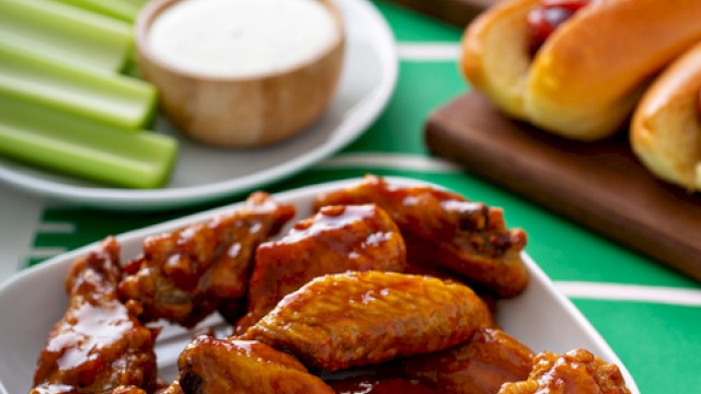Buffalo chicken wings with celery sticks, ranch dipping sauce, potato chips, onion rings, hot dogs, and chocolate brownies on a football-themed table.