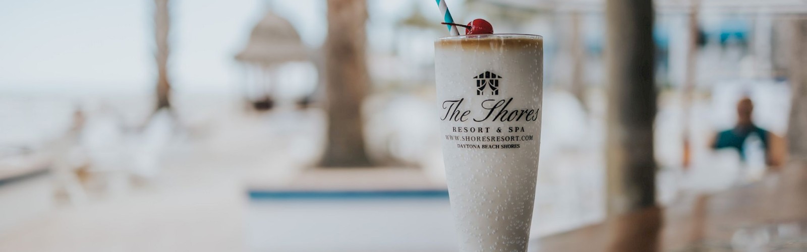 A frosty beverage with a cherry on top and a straw, placed on a wooden surface at a beachside resort with palm trees in the background.