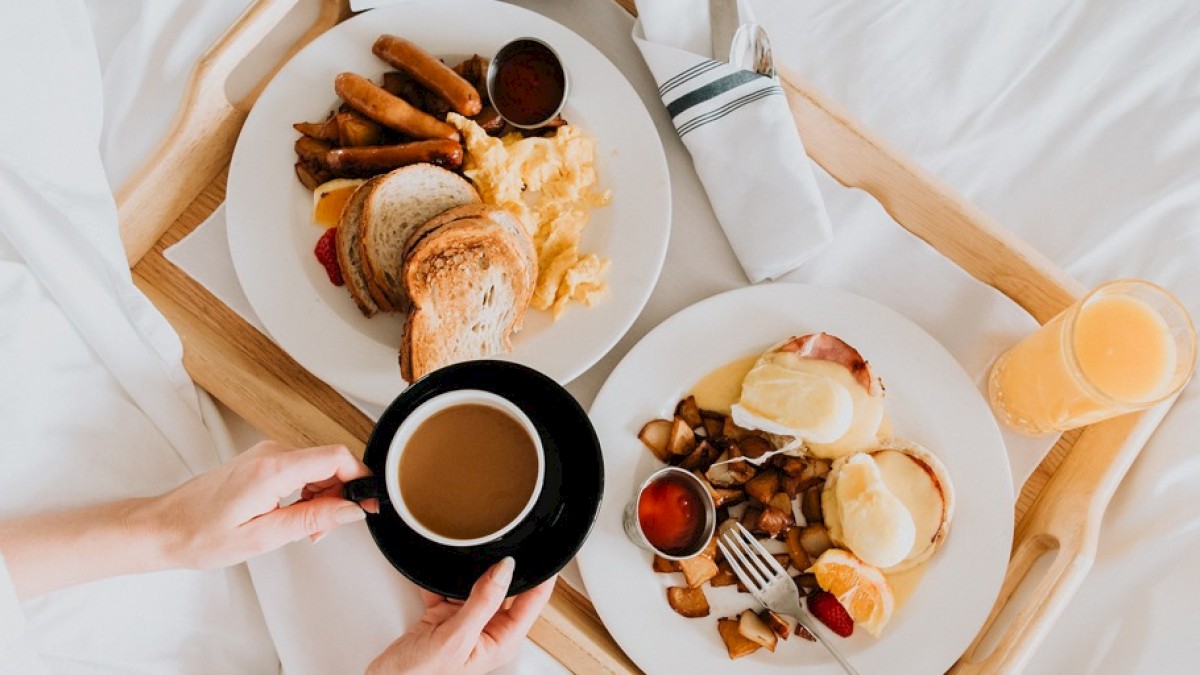 Two plates of breakfast with toast, sausages, scrambled eggs, eggs benedict, diced potatoes, a cup of coffee, and a glass of orange juice on a tray.