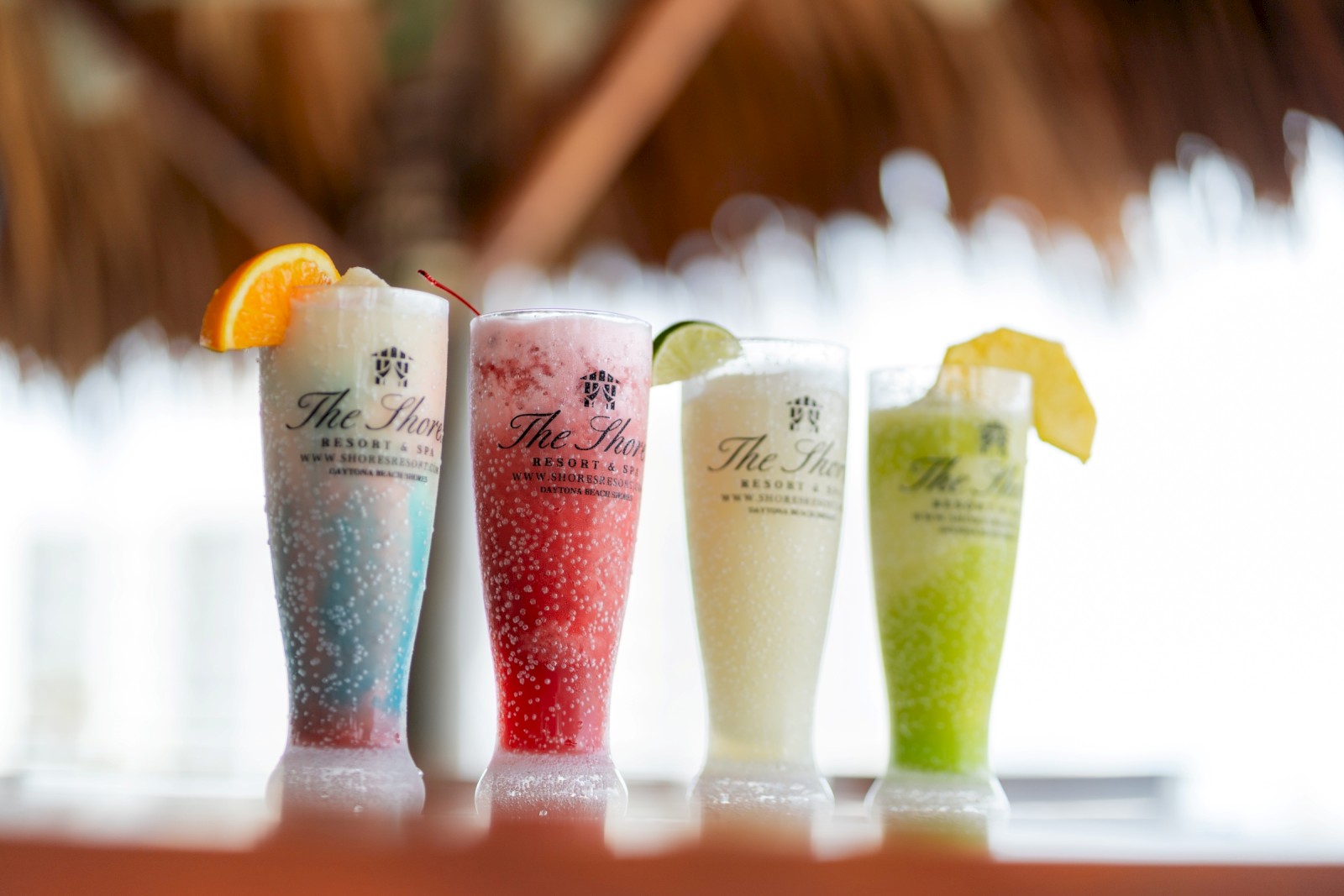 Four colorful tropical drinks with fruit garnishes are lined up on a table under a thatched roof at a resort.