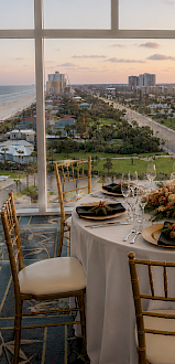 A beautifully set round dining table with elegant black napkins and gold plates, overlooking a sunset view from a high-rise with beach and cityscape.