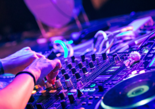 A person is using DJ equipment with colorful lights in a lively setting, adjusting knobs and preparing music mixes for an event.