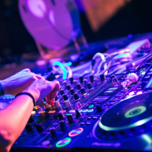 A person is using DJ equipment with colorful lights in a lively setting, adjusting knobs and preparing music mixes for an event.