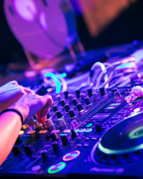 A person is using DJ equipment with colorful lights in a lively setting, adjusting knobs and preparing music mixes for an event.