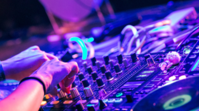 A person is using DJ equipment with colorful lights in a lively setting, adjusting knobs and preparing music mixes for an event.