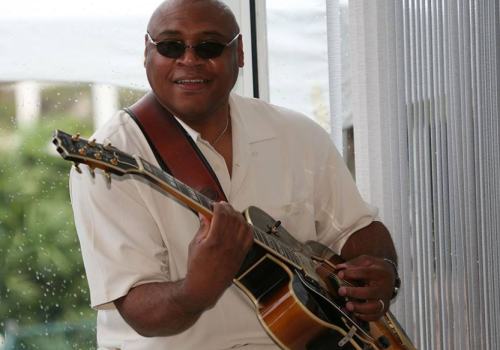 A person wearing sunglasses and a white shirt, playing an acoustic guitar, is smiling indoors with a window in the background.