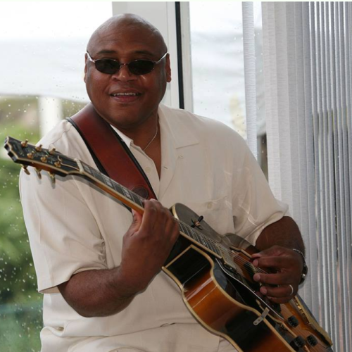 A person wearing sunglasses and a white shirt, playing an acoustic guitar, is smiling indoors with a window in the background.