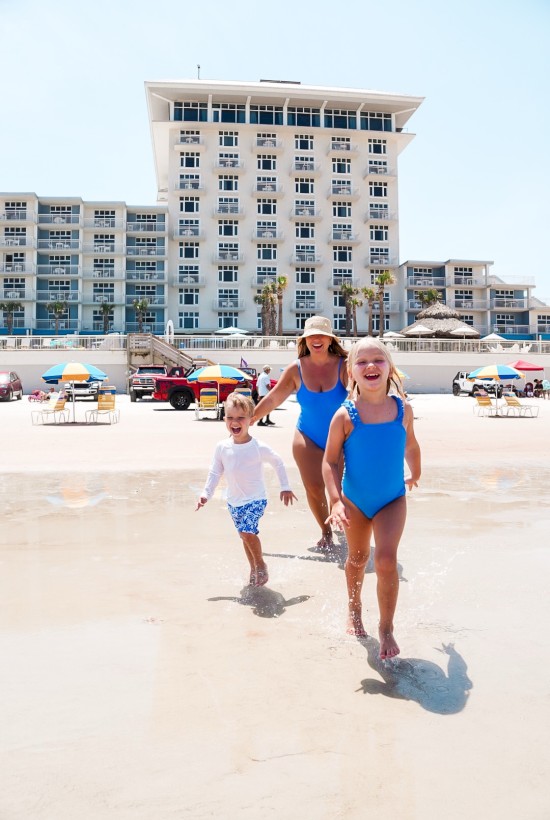 A woman and two children walk joyfully on a sunny beach with a large hotel and umbrellas in the background.
