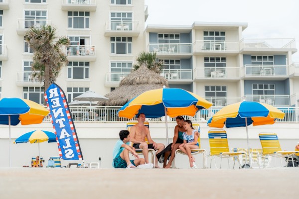 A group of four people enjoying a sunny day at the beach, sitting under colorful umbrellas with a hotel in the background.