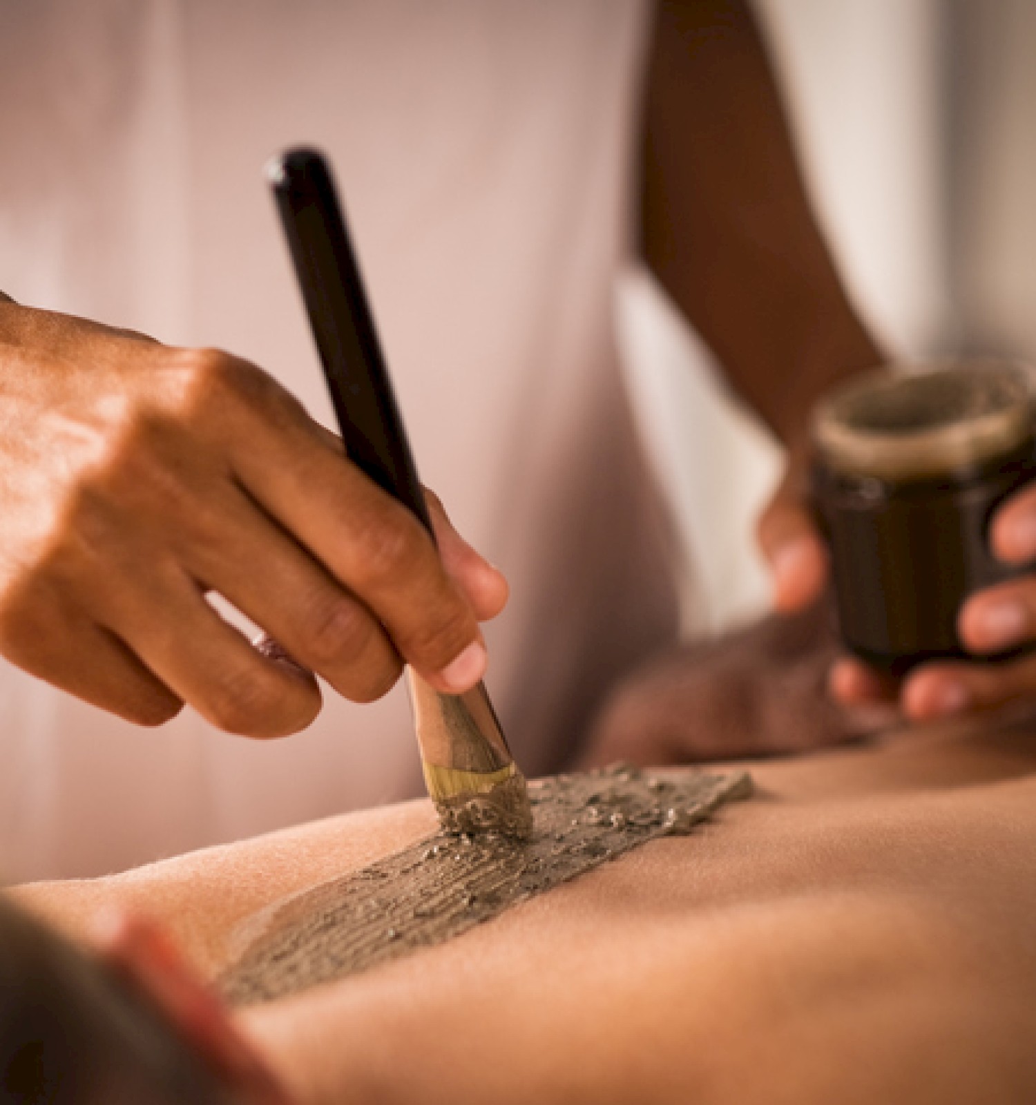 A person is applying a mud mask or scrub to someone’s back using a brush while holding a container, likely at a spa or wellness center.