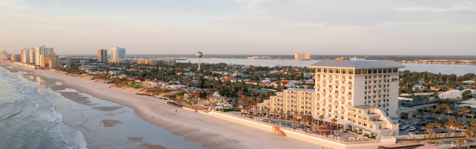 A coastal beach scene shows sandy shores, ocean waves, and a cityscape with several buildings under a clear sky.